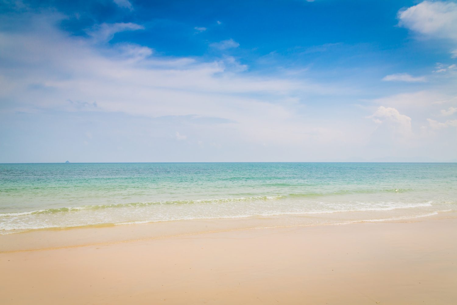 Tranquil beach with soft waves lapping against the shore under a bright blue sky with scattered clouds.