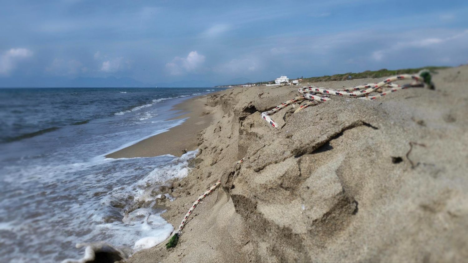 A coastal erosion scene showing a sandy shoreline with visible cliff-like edges formed by waves, and a rope lying on the sand.
