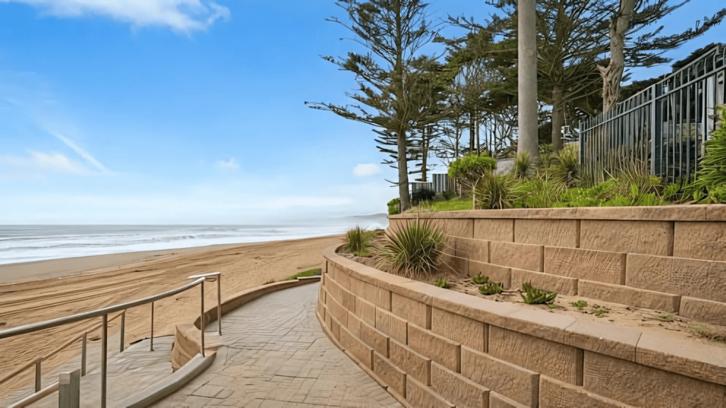 Curved stone retaining wall with plants overlooking a sandy beach, a paved path, and the ocean under a clear blue sky.