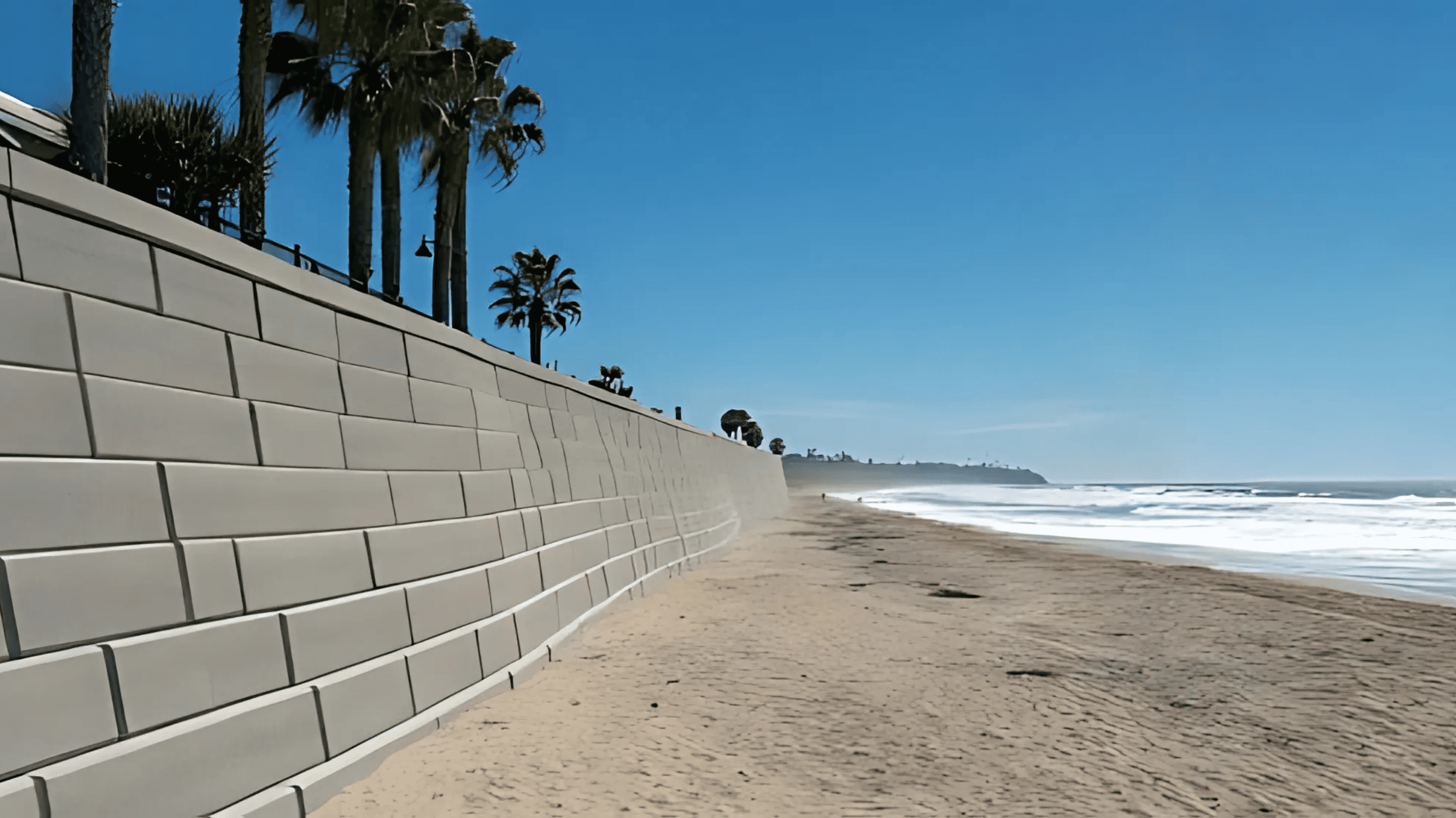 Smooth concrete retaining wall along a wide sandy beach with palm trees above, overlooking ocean waves under a bright blue sky.