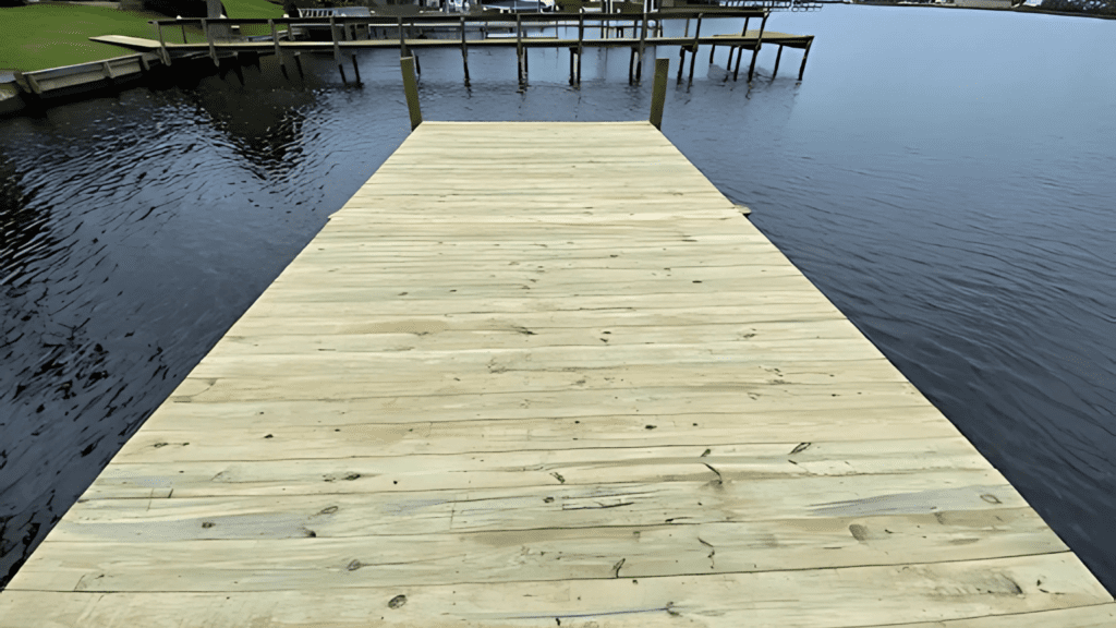 Wooden dock extending over calm water, surrounded by other docks, grassy lawns, and houses under a cloudy sky.