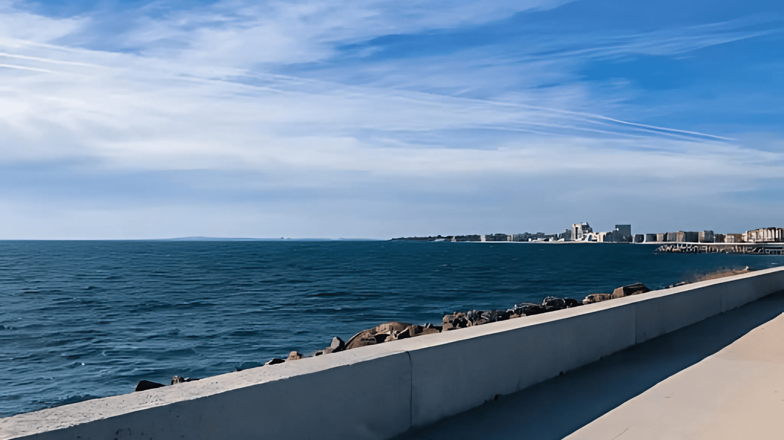 Concrete seawall overlooking calm blue ocean waves, with distant buildings and a clear sky in the background.