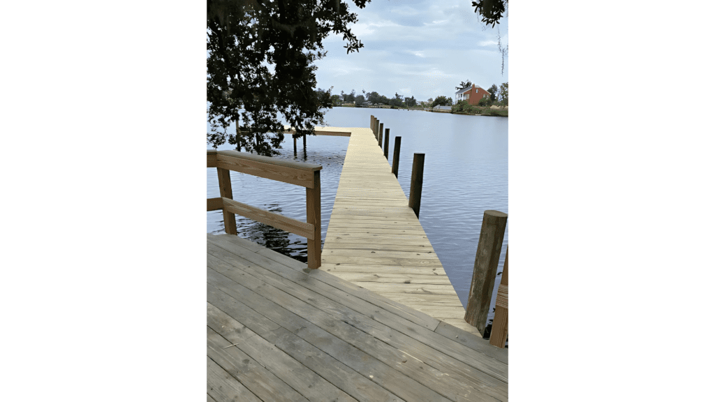 Wooden boat dock extending over calm water, surrounded by trees and a red house in the distance under a cloudy sky.