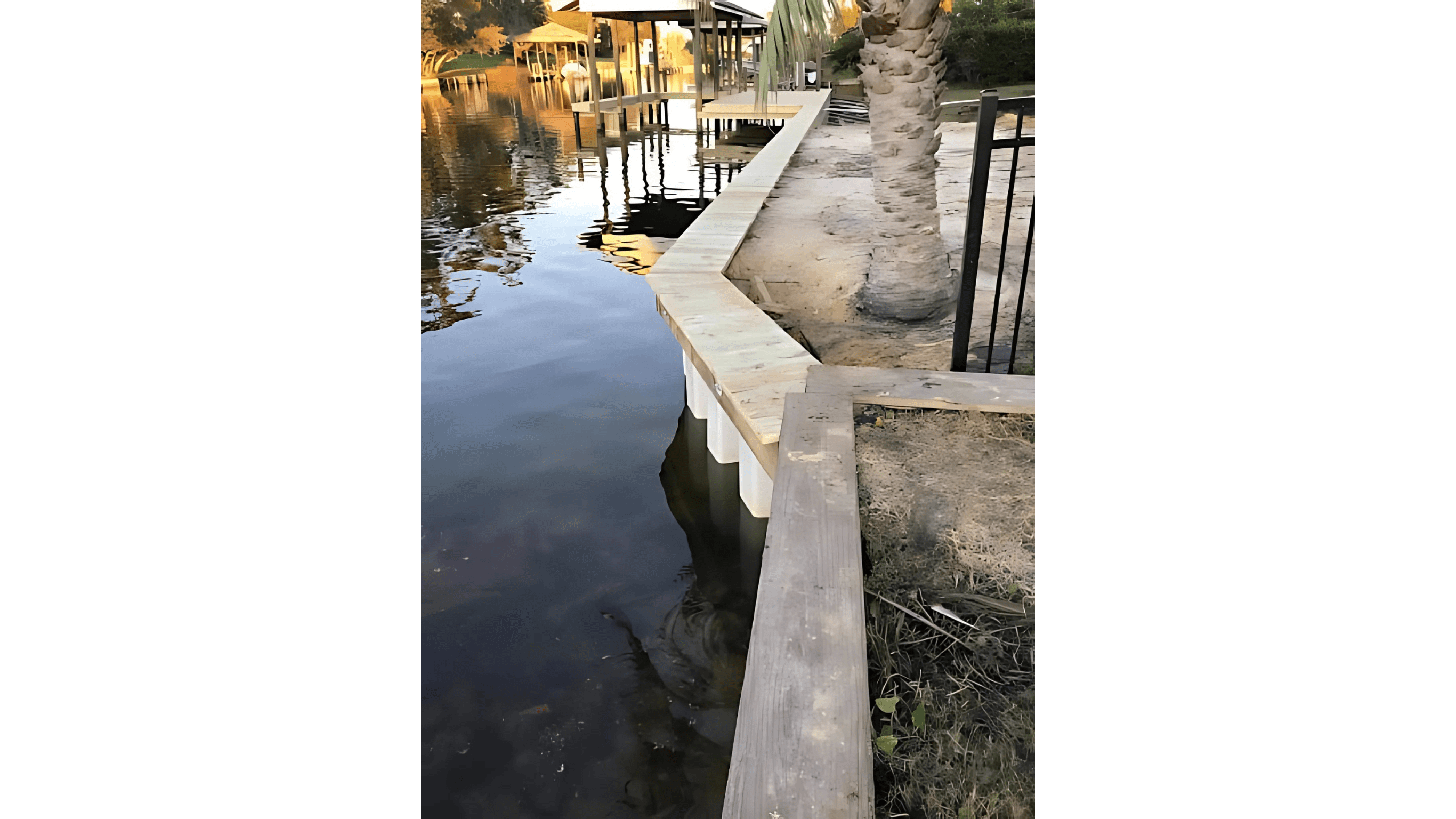 Vinyl seawall with a wooden cap along a calm waterfront, bordered by sand, grass, a palm tree, and nearby docks.