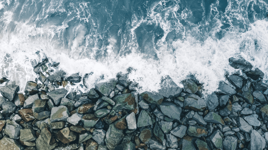 Aerial view of ocean waves crashing against large rocks placed as a shoreline barrier, resembling a bulkhead or seawall.