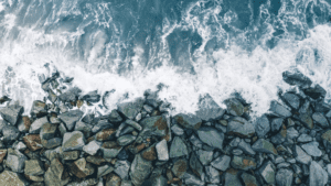 Aerial view of ocean waves crashing against large rocks placed as a shoreline barrier, resembling a bulkhead or seawall.