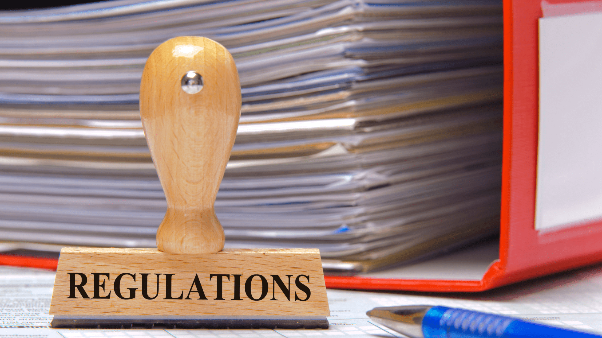 A wooden stamp labeled "REGULATIONS" sits on a desk with stacked documents, a red binder, and a blue pen, symbolizing compliance.