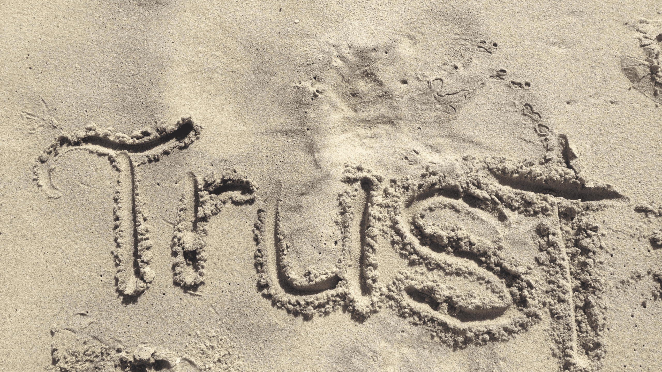 The word "Trust" is written in the sand with footprints nearby, creating a temporary message on the beach before the tide washes it away.