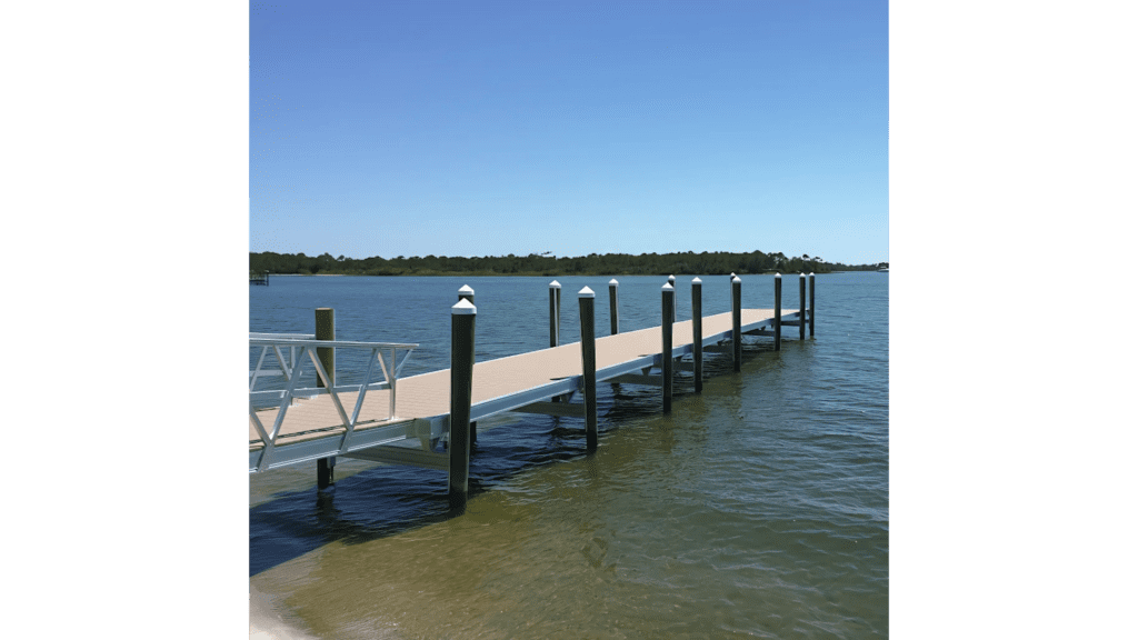 A long wooden dock with white post caps extends over calm, clear water, with a metal gangway leading from the sandy shore to the pier.
