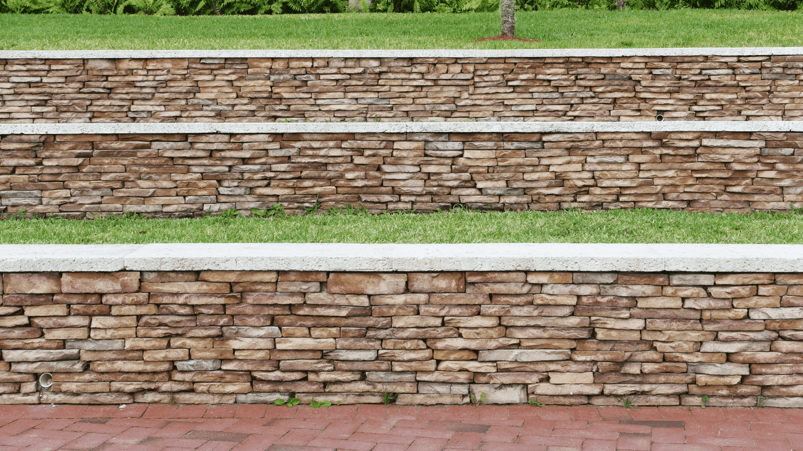 Three tiers of stacked stone retaining walls with white concrete caps and grass in between, built against a gentle green slope.
