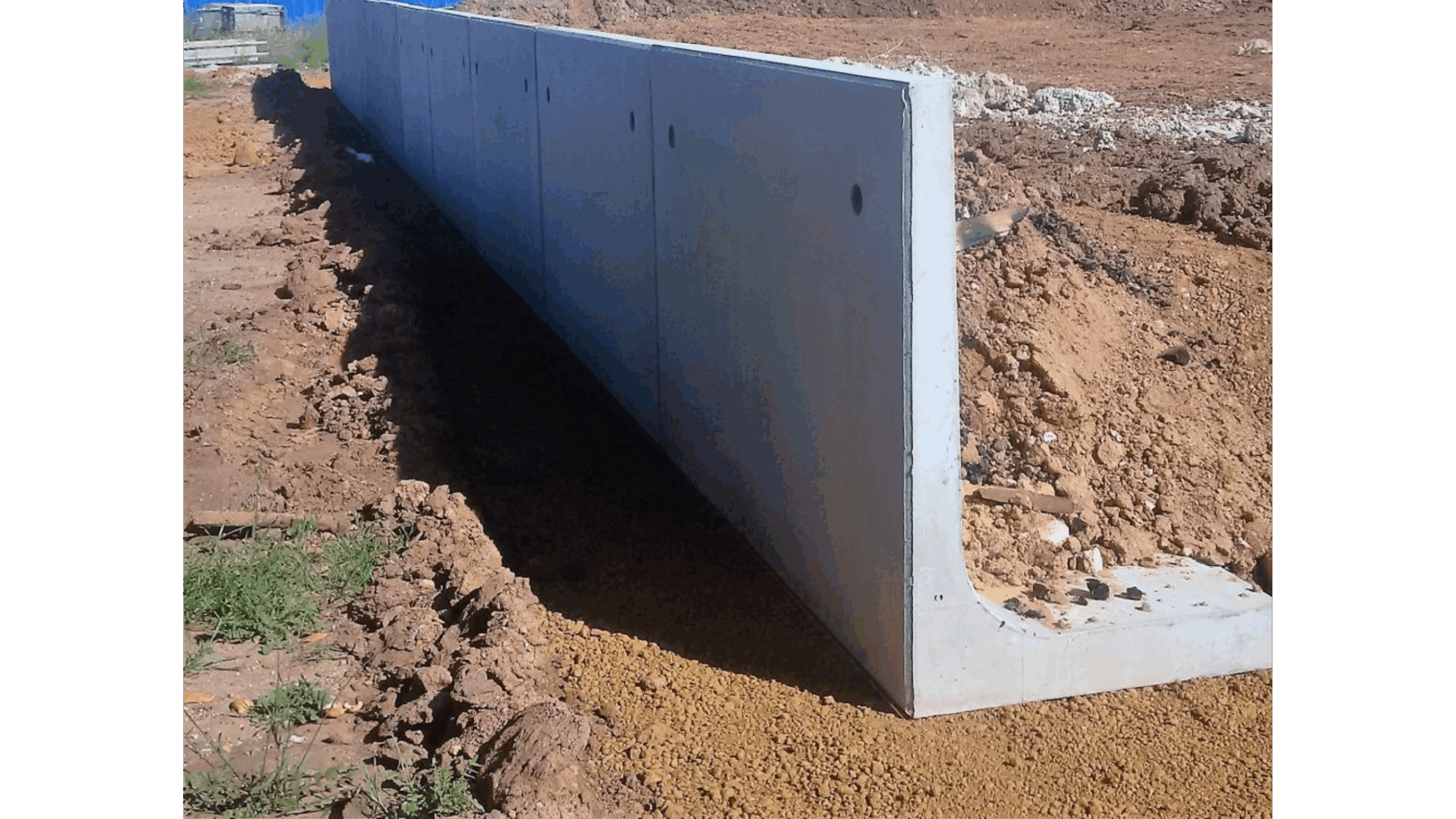 L-shaped concrete cantilever retaining wall under construction, with compacted soil behind and an exposed footing on a dirt site.