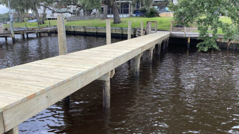 A newly constructed wooden dock extends over calm water, supported by sturdy pilings, with a grassy shoreline and trees in the background.