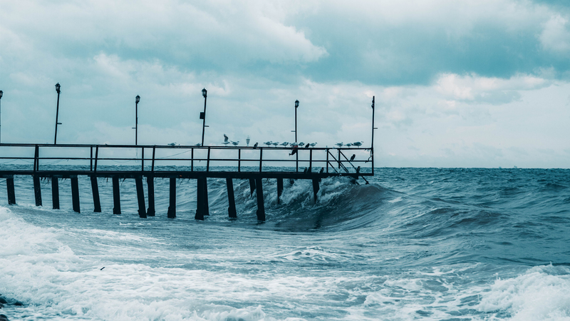 A weathered pier with leaning lampposts stands over rough ocean waves, as seagulls perch on the railing under a cloudy sky.