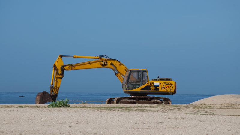 A yellow excavator with graffiti sits on a sandy shore near the ocean, its bucket resting on the ground, used for marine construction.